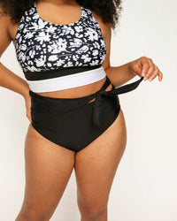 A studio image of a women pointing out the tie detail on black swimsuit bottoms.