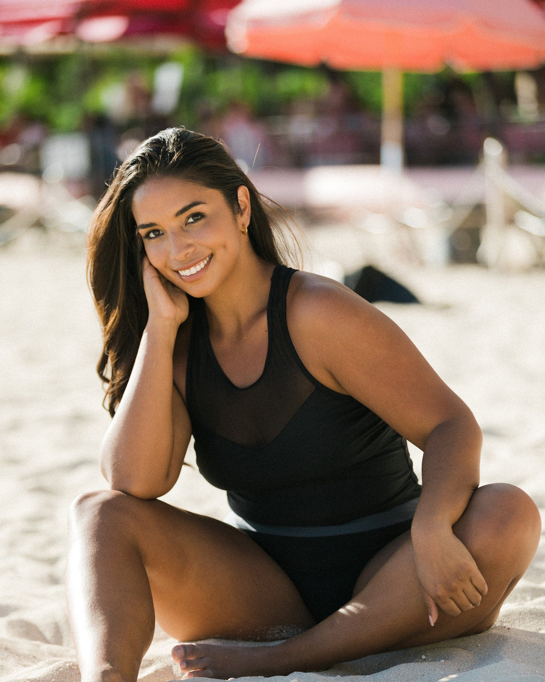 A women sitting at the beach wearing an athletic black swimsuit top with mesh detailing.